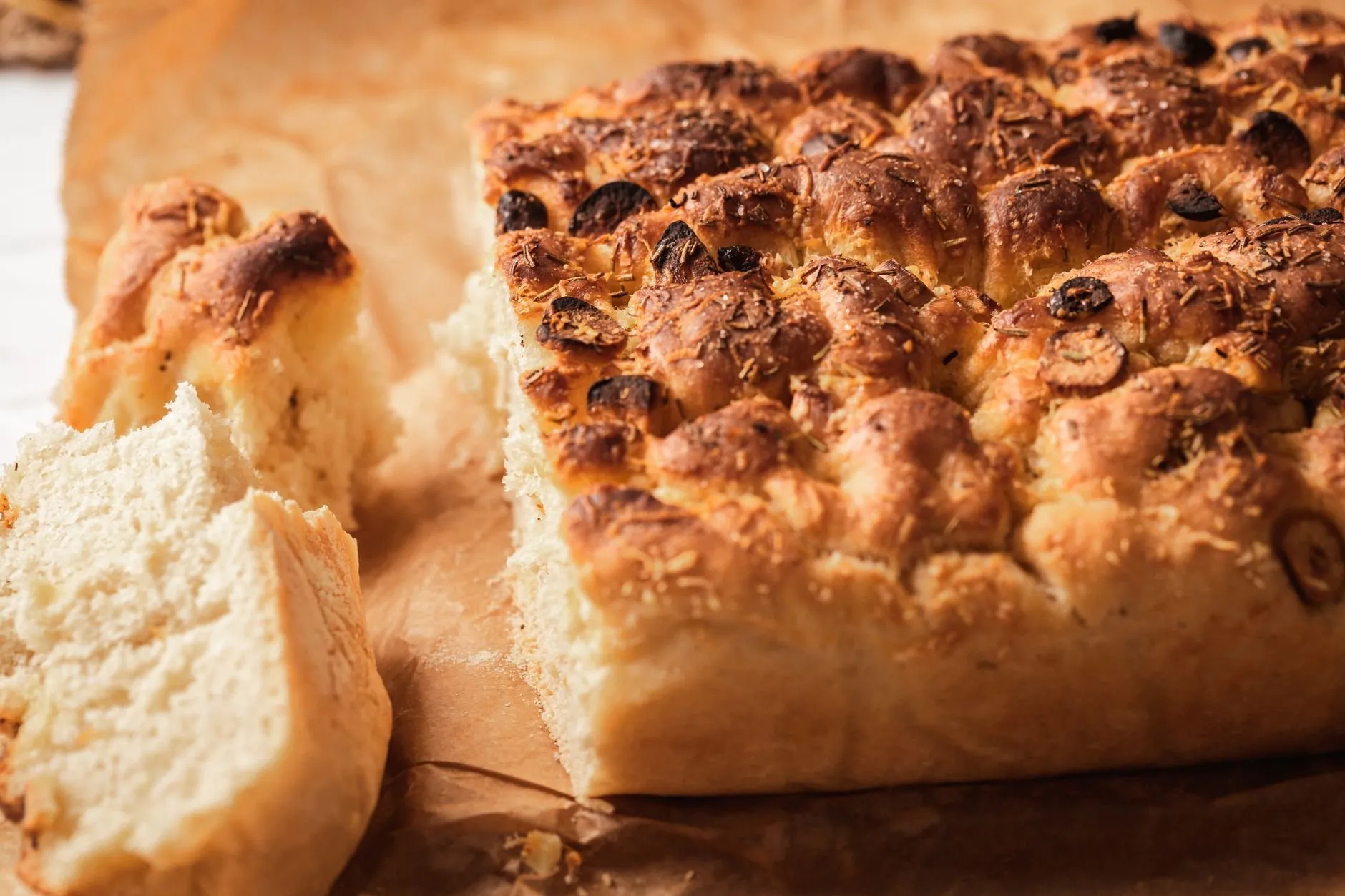 A slab of golden focaccia on baking paper with a torn piece showing its soft crumb